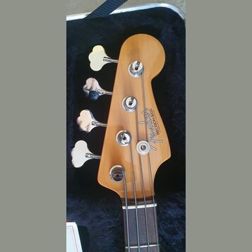 Hlava čtyřstrunné basové kytary z přírodního dřeva v černém kufru. Na hlavě je vidět logo Fender a nápis PRECISION BASS, pod kterým je drobný nečitel…
