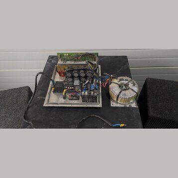 Fotografie zachycuje vnitřek elektronického zařízení, pravděpodobně zesilovače, bez krycího šasi. Je vidět hliníkový základ s instalovanými kondenzát…