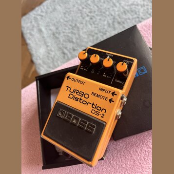 Oranžový kytarový efekt s černým spodním panelem a ovládacími knoflíky. Na horní straně je nápis TURBO Distortion DS-2 a v dolní části logo BOSS. Jso…