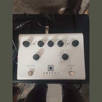 Bílý kytarový pedál s nápisem AMPED 1 a pod ním menším textem DEPT. 10 | 100W AMP PEDAL. Nad nápisem je logo ve tvaru hvězdy v černém čtverci. Zaříze…