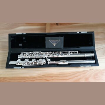 Stříbrná flétna v černém pevném kufru. Na krabičce uvnitř kufru je nápis SANKYO FLUTES, MFG. CO., LTD. JAPAN a pod ním trojúhelníkové logo. Nástroj v…