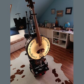 Na fotografii je hybridní nástroj kombinující kontrabas a buben. Na bílém bubnovém povrchu je černý nápis „Country music session Kontrabass Banjo“. N…