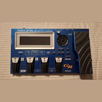 Modrý kytarový syntetizátor s nápisem Roland GR-55 GUITAR SYNTHESIZER. Zařízení má displej, ovládací knoflík se šipkami a čtyřistupňové pedály označe…