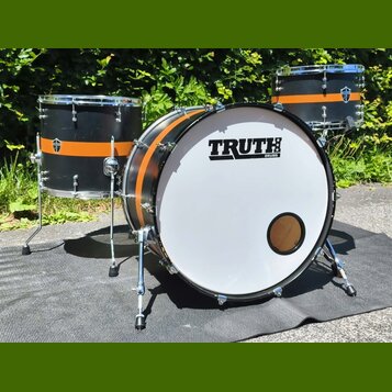 Černá bubnová sada s oranžovými pruhy, která obsahuje basový buben a dva tom-tomy. Na bílém předním zrcadle basového bubnu je černý nápis TRUTH DRUMS…