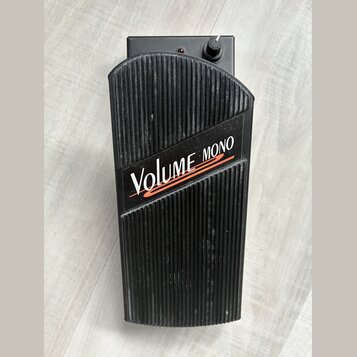 Černý volume pedál s žebrovaným povrchem. Na horní části je bílý nápis VOLUME a červený nápis MONO, pod kterými jsou dvě červené vlnovky. V horní čás…