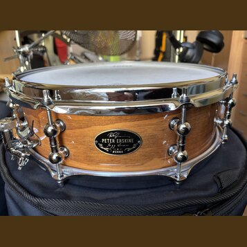 Dřevěný buben s přirozeným povrchem a chromovanými obručemi. Na korpusu je oválný černý štítek s nápisy „PETER ERSKINE“, „Jazz Snare“ a logem „TAMA“.…