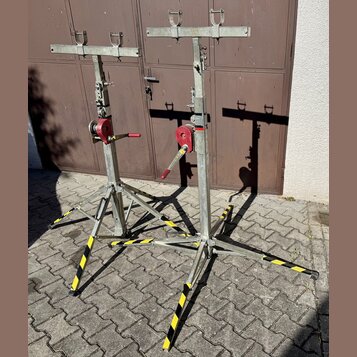 Dvě kovové stojany s nastavitelnou výškou a horními popružími pro uchycení nástroje. Stojany mají stříbrný kovový povrch, červené mechanismy pro nast…