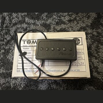 Černý humbucker s šesti viditelnými šrouby v polosovém uspořádání. Na povrchu je rukopisní nápis Tuma. Snímač má připojený černý kabel a leží na bílé…