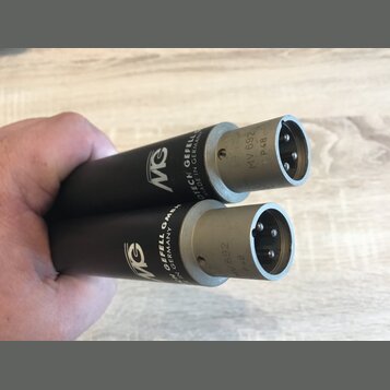 Dvě černé válcové součástky s kovovými konektory na koncích. Na tělech je bílé logo MG a nápisy GEFELL GMBH, MADE IN GERMANY. Na kovových částech je…