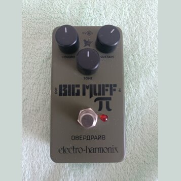 Šedý kytarový efekt s nápisy BIG MUFF π, ОВЕРДРАЙВ a electro-harmonix. Na horní straně jsou tři černé otočné knoflíky s popisky SUSTAIN, TONE a VOLUM…