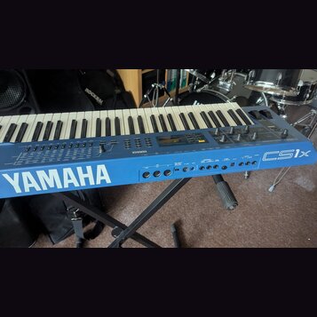 Modrý synthesizér s klaviatura umístěný na černém stojanu. Na přední straně je bílý nápis YAMAHA a vpravo označení modelu CS1x. Na ovládacím panelu j…