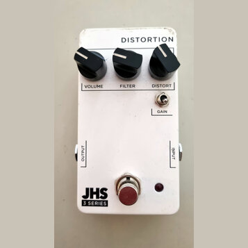 Bílý kytarový efekt s nápisem DISTORTION. Na horní straně jsou tři černé otočné ovnovky s popisky VOLUME, FILTER a DISTORT, dále je zde přepínač GAIN…