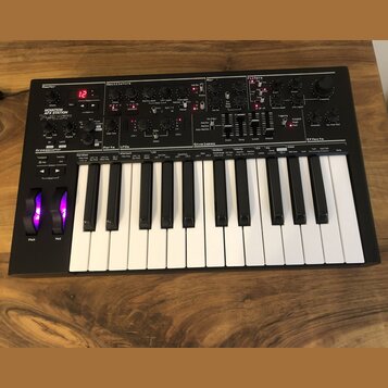 Černý syntetizátor s klaviaturaí a ovládacími prvky v horní části. Na levé straně je nápis NOVATION AFTERTOUCH XL. Přístroj obsahuje početné otočné k…