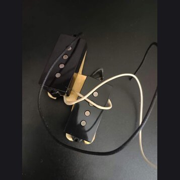 Dvě elektrické kytarové snímače typu humbucker s černými plastovými kryty a viditelnými polkovými magnety. Snímače jsou umístěny na světlém dřevěném…