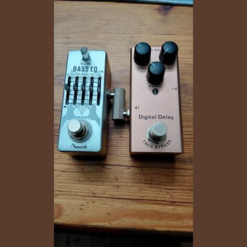 Dvě efektové pedály umístěné vedle sebe na dřevěném povrchu. Levý pedal je stříbrný s nápisy VOLUME, BASS EQ a značkou Amuzik. Pravý pedal je růžozla…