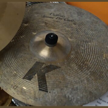 Zlatý cimbál s koncentrickými rýhami na povrchu. V horní části je nápis „Custom Special Dir Crash“, ve spodní části je velké černé písmeno „K“. Povrc…