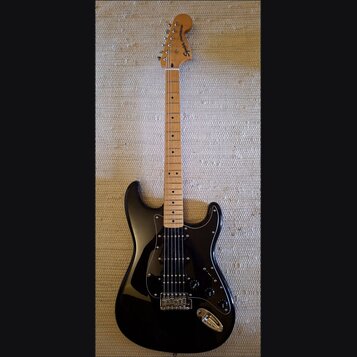 Černá elektrická kytara s jasným dřevěným hmatníkem a bílým pickguardem. Na hlavě je nápis Squier STRATOCASTER. Kytara má tři snímače, pětipositionní…