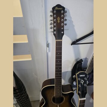 Hnědá akustická kytara s 12 strunami. Na hlavě je zlatý nápis Ibanez a pod ním zlatý symbol šipky. Kytara má tmavý hnědý povrch s výrazným lemováním…