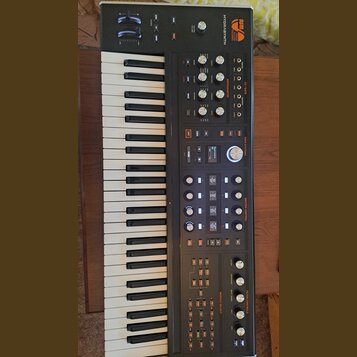 Černý syntetizátor s klaviaturaí. Na horním panelu je oranžové logo ASM Electric Music Company a nápis HYDRASYNTH. Přístroj obsahuje numeroustovací k…