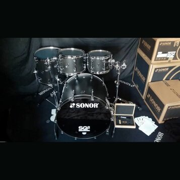 Černá bicí souprava značky SONOR, model SQ2. Sestava obsahuje basový buben s nápisem SONOR a SQ2, dva tom-tomy a jeden floor tom. Vedle nástrojů jsou…