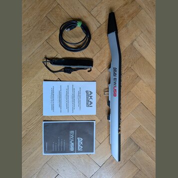 Na fotografii je zobrazen MIDI kontrolér AKAI EWUSB v šedé barvě s černými detaily. K nástroji patří USB kabel, popruh a dva printed manuály s nápisy…