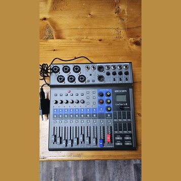 Šedý digitální mixážní pult s nápisem ZOOM LiveTrak L-8. Zařízení obsahuje několik vstupních konektorů, posuvníky, tlačítka a malý displej. Povrch je…