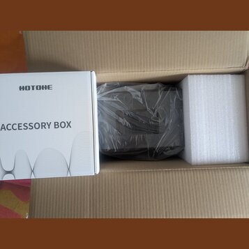 V hnědé kartonové krabici se nachází bílé papírové balení s nápisy HOTONE a ACCESSORY BOX. Vedle něj je tmavý, pravděpodobně plastový předmět zabalen…