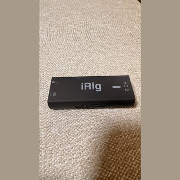 Černý obdélníkový audio rozhraní s nápisem iRig a označením HD 2. Na levé straně je vidět symbol mikrofonu a ikona baterie. Povrch zařízení je matný,…