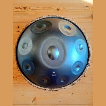 Kulatý kovový handpan s modravým, gradientním povrchem a několika vyhlubecinami (tóny). Nástroj má pletený lem v béžové barvě. V dolní části je bílý…
