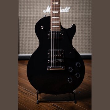 Černá elektrická kytara s tvarem single-cutaway a dvěma humbuckery. Na hrazdiku v pozadí je vidět bílý nápis Marshall. Kytara má čtyři ovládací knofl…