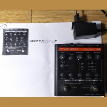 Černý efektový pedál s oranžovým pruhem, na kterém je nápis TC-HELICON harmonyM a VOICETONE™. Zařízení obsahuje několik otočných potenciometrů, tlačí…