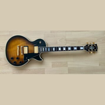 Elektrická kytara s tónem sunburst a černým povrchem na okrajích. Na hlavě je logo Gibson a nápis Les Paul Custom. Nástroj má zlatý hardware, dva hum…