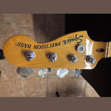 Fotografie zachycuje hlavu basové kytary z přírodního dřeva s čtyřmi chromovanými ladičkami. Na povrchu je černý nápis Squier BY FENDER a PRECISION B…