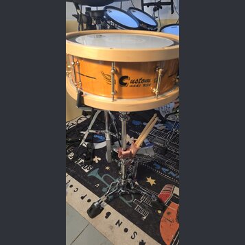 Dřevěný buben v přírodním odstínu s nápisem Custom made USA. Nástroj je umístěn na chromovaném stojanu, ke kterému je připevněn růžový držák dvou bub…