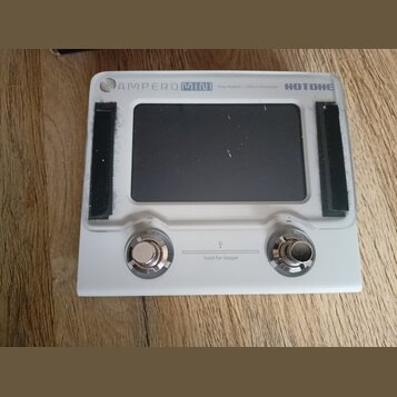 Bílý efektový procesor s černým displejem. V horní části je nápis AMPERO MINI Amp Modeler / Effects Processor a logo HOTONE. Dole jsou dvě kovové kon…