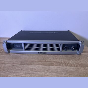 Stříbrný rackový výkonový zesilovač s černým horním krytem. Na předním panelu je nápis PLX 1804 PROFESSIONAL POWER AMPLIFIER a v dolní části logo QSC…