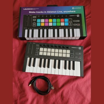 Černý MIDI kontrolér Novation Launchkey Mini s bílými a černými klávesami. Na horní části jsou umístěny šedé pady, ovládací knoflíky a nápis LAUNCHKE…