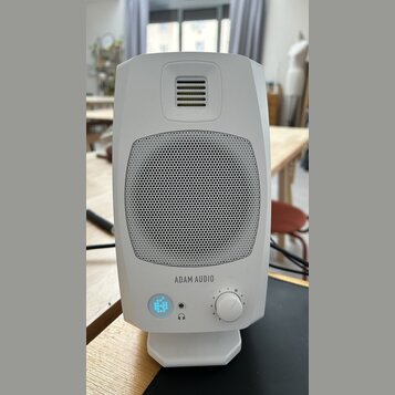 Bílý studiový monitor značky ADAM AUDIO s kovovou mřížkou reproduktoru. V dolní části jsou umístěny ovládací prvky: podsvícené tlačítko s ikonou, vst…