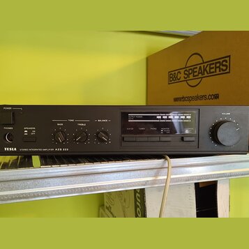 Černý stereofonní zesilovač s nápisem TESLA STEREO INTEGRATED AMPLIFIER AZS 223. Na předním panelu jsou ovládací prvky pro basy, treble a balance, př…