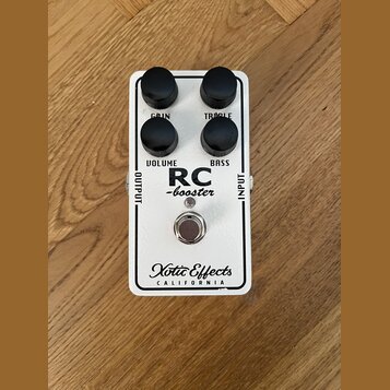 Bílý kytarový efekt s nápisy RC -booster a Xotic Effects CALIFORNIA. Na horní straně jsou čtyři černé otočné knoflíky s popisky GAIN, TREBLE, VOLUME…