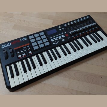 Černý MIDI kontrolér s klaviaturní součástí a bílými a černými klávesami. Na levé straně je nápis AKAI professional MPK49 a mřížka šedých padů. Horní…