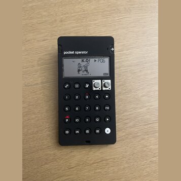 Černý přenosný syntezátor s nápisem pocket operator. Displej zobrazuje grafiku dvou postav, text K.O! a označení PO6. Zařízení má numerickou klávesni…