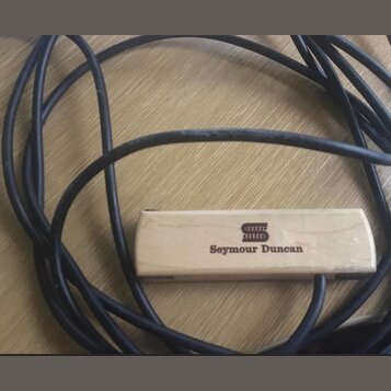 Na fotografii je vidět černý kabel a k němu připevněný obdélníkový dřevěný blok světlé barvy. Na bloku je vyryto logo a nápis Seymour Duncan. Povrch…