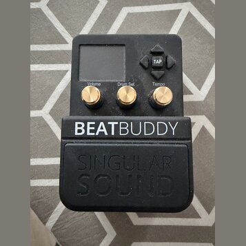Černý efekt pedál s nápisem BEATBUDDY a pod ním vylisovaným logem SINGULAR SOUND. Zařízení má malý displej, čtyři šipkové tlačítka s nápisem TAP a tř…