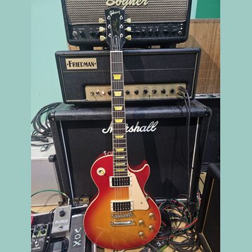 Elektrická kytara s tvarem singlecut a povrchovou úpravou sunburst v červeně a žluté. Na hlavě je bílé logo Gibson a nápis Les Paul Custom. Kytara má…