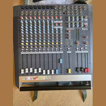 Šedý mixážní pult s nápisem ALLEN&HEATH a označením modelu PA12. Zařízení obsahuje řadu vertikálních faderů, barevné otočné knoflíky a různé typy vst…