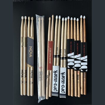 Sada různých bubnových paliček z přírodního dřeva rozložená na černém povrchu. Jsou zde paličky s nápisy NOVA, Vincent, Pro-Mark a Vic Firth (označen…