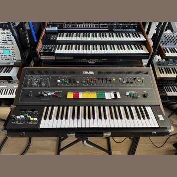 Hnědý syntetizátor značky YAMAHA s klaviatura a ovládacím panelem s různobarevnými tlačítky. Nástroj je umístěn v černém přepravním kufru a stojí na…