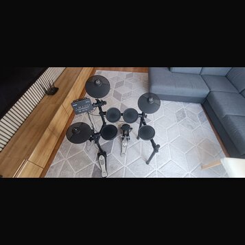 Elektronická bicí souprava s černými padmi a činely na kovovém stojanu. Na ovládacím modulu je vidět nápis YAMAHA a označení DTX. Souprava obsahuje č…