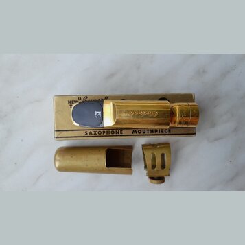 Zlatý mosazný saxofonový mouthpiece s černým náústkem, položený na kartónové krabičce. Na mouthpiece je vyryt nápis "Otto Link". Krabička obsahuje te…